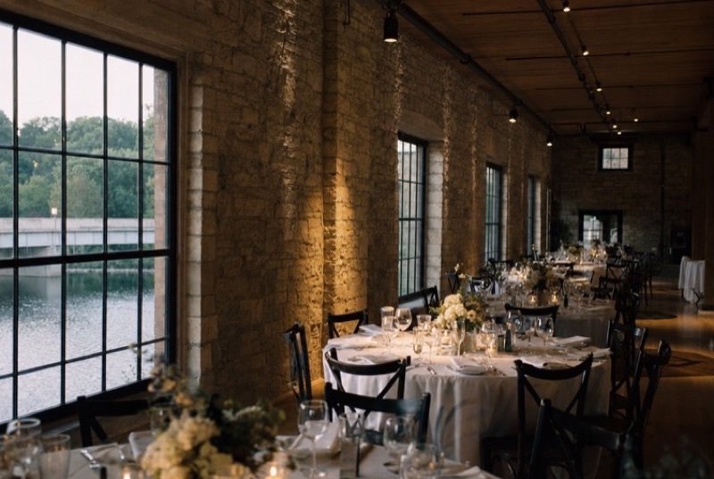 A long hall with stone walls and tall windows, round tables dressed in white linen with flowers and candles for a dinner