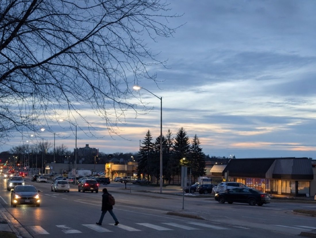 A suburban commercial road at dusk lined with single-storey plazas of small shops, car headlights on and a pedestrian at a crosswalk