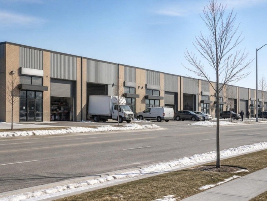 A row of small brick and metal-panel units with roll-up doors in a business park on a cold spring morning, vans parked in front
